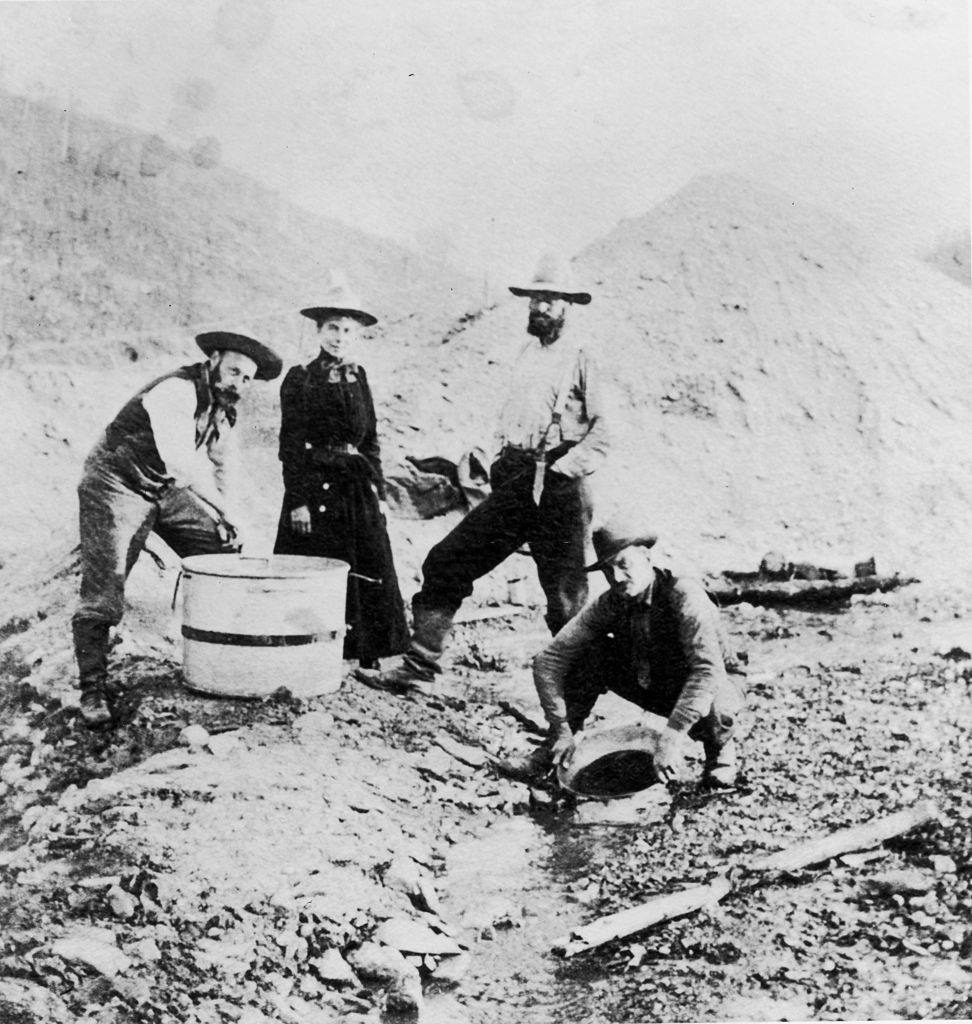 Three men and a woman standing and crouching around a riverbed while panning for gold during the Klondike gold rush, Alaska, 1895.