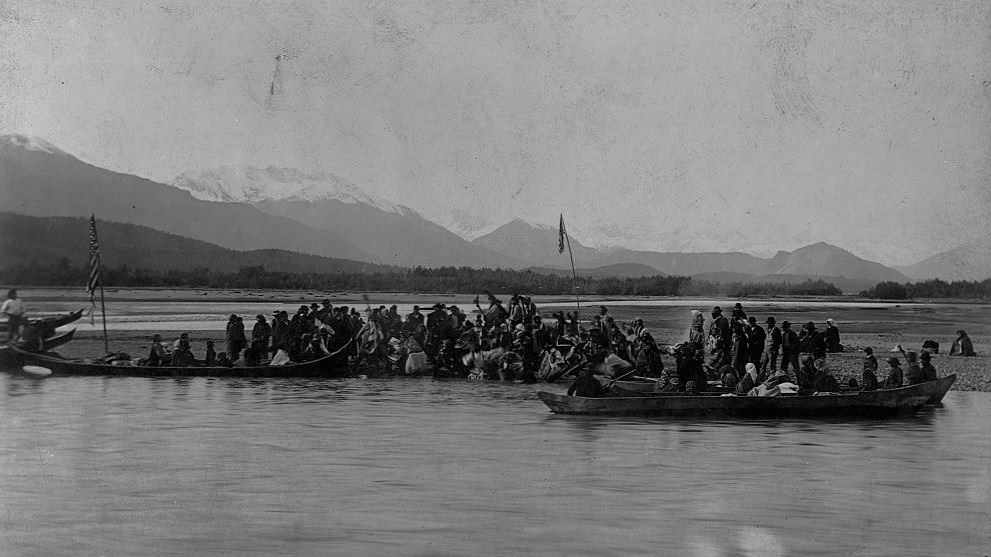Tlingits gather on bank of a gravely sandspit for a potlatch dance, prior to the actual potlatch feast, along the Chilkat River. Alaska 1895.