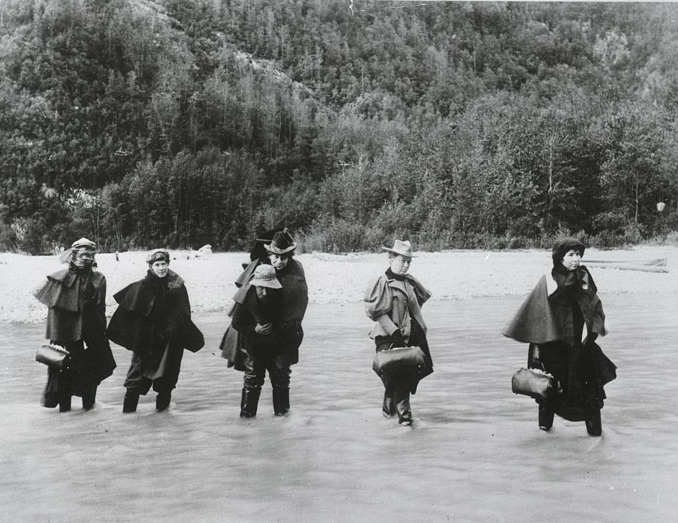 A man carries an actress across an Alaskan river while others must ford it to get to a gold mine, 1897.