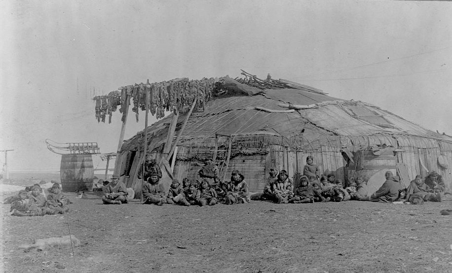 A group of Eskimos sit out side a large round house build out of wood and animal skins. Walrus meat dries on poles next to entrance, St. Lawerence Island, Alaska, 1897.