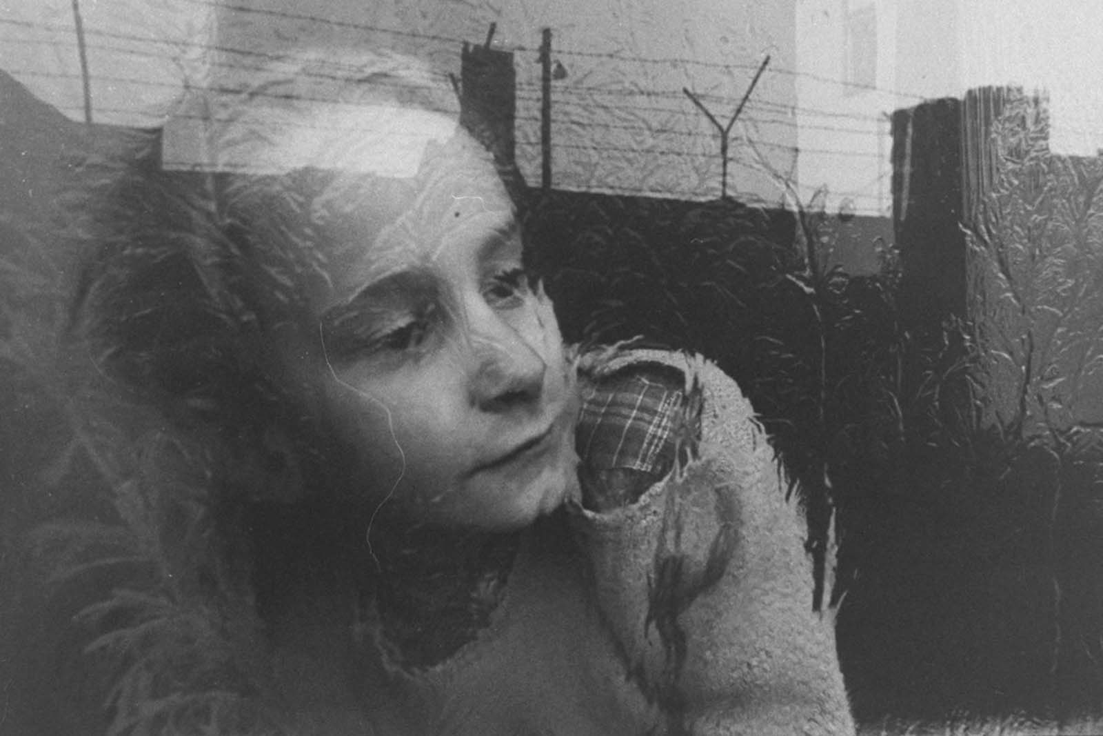 A young girl gazes pensively through the pane of her apartment window, which reflects the image of barbed wire fencing that tops the nearby Berlin Wall, in December 1962.