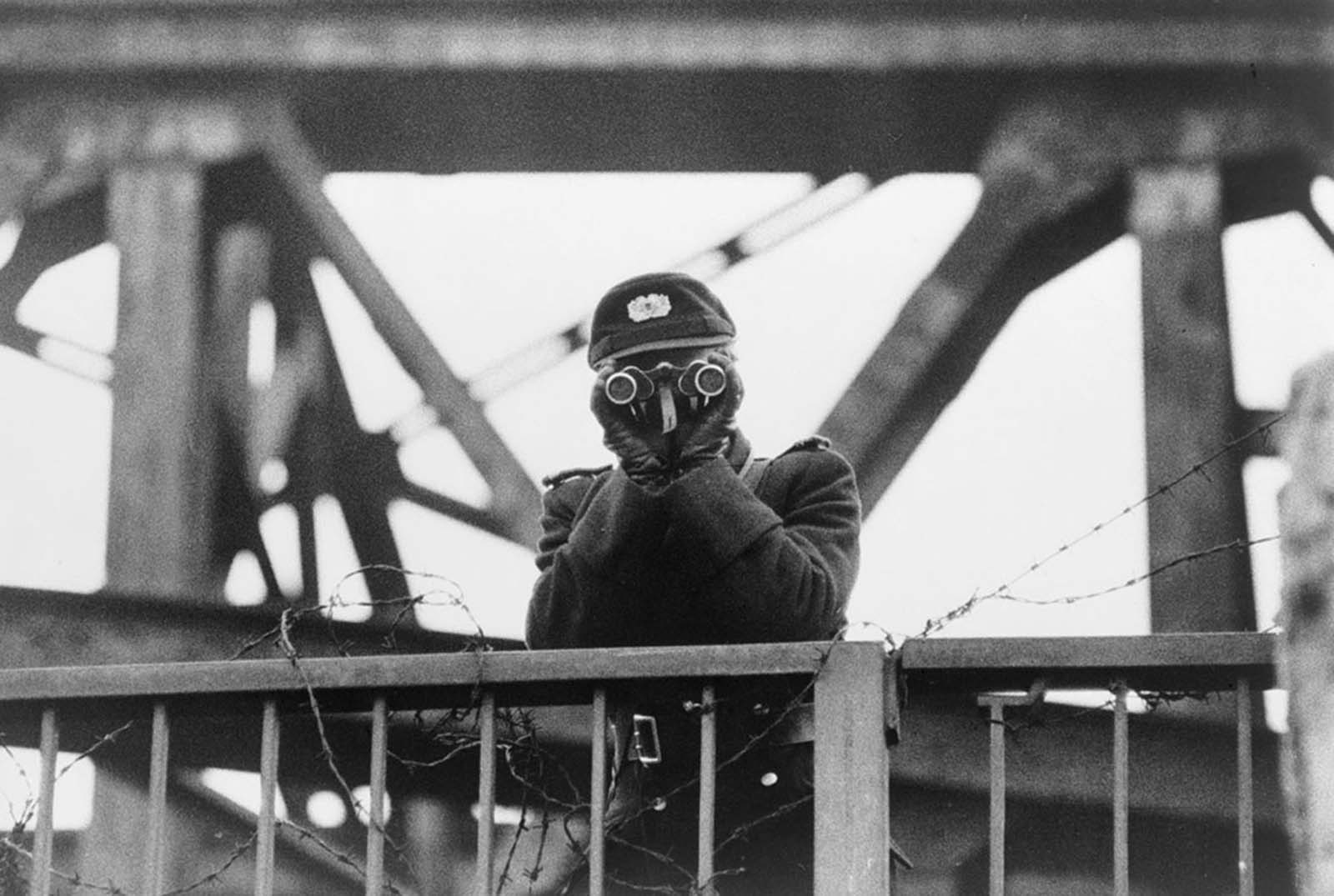 An East German VOPO (Volkspolizei) border policeman uses binoculars while standing guard on one of the bridges linking East and West Berlin, in 1961.