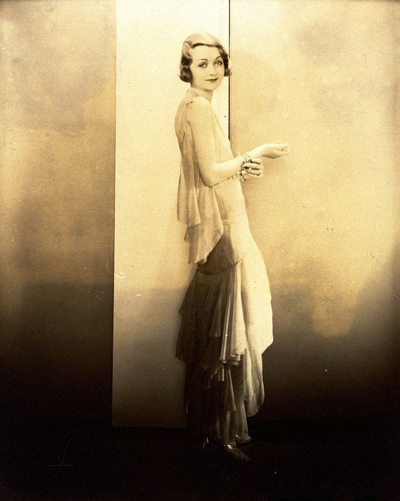 Constance Bennett wearing sleeveless chiffon gown with back scarf and flounces that spiral down the skirt, Vogue 1929.