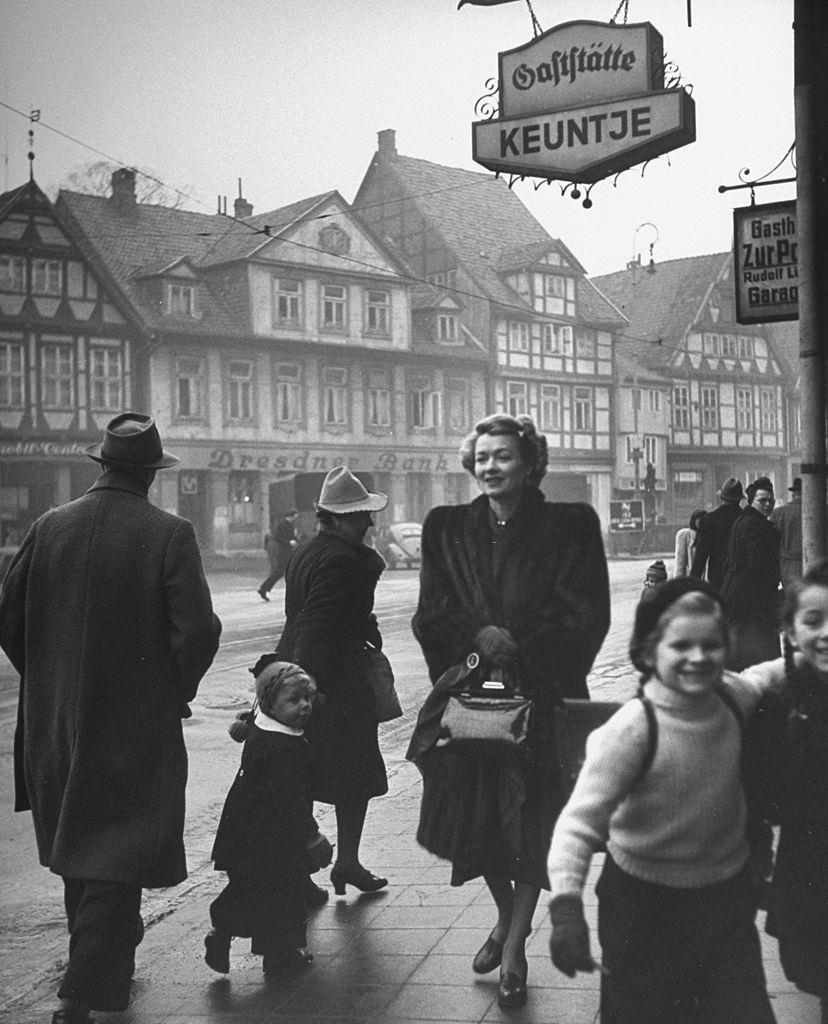 Constance Bennett walking down a shop lined street.