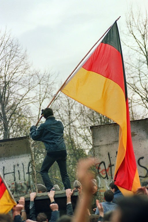 A West Berliner prepares to hand over a FRG flag to East German Vopos through a portion of the fallen Berlin Wall near the Brandenbourg Gate early 11 November 1989.