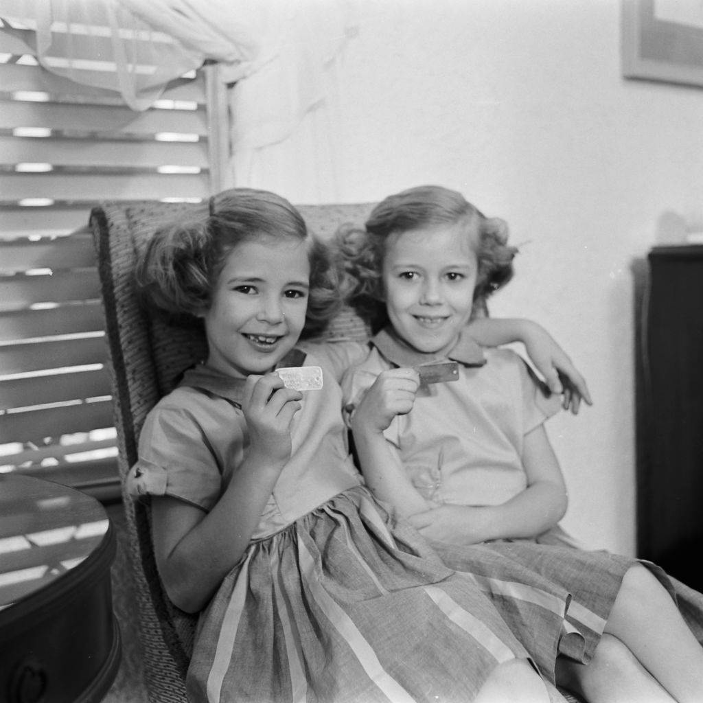 Two sisters sitting in a chair in their home after an atomic war drill with their family, holding up the identification tags they wear around their necks, March 1954.