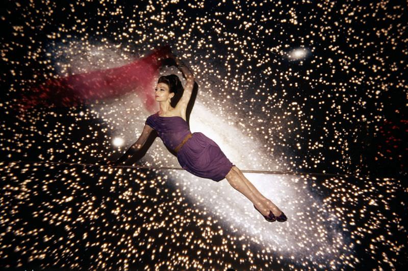 Floating against a backdrop of the galaxy, Isabella is wearing violet short evening dress with pleats which drape across one shoulder by Maurice Rentner, 1960