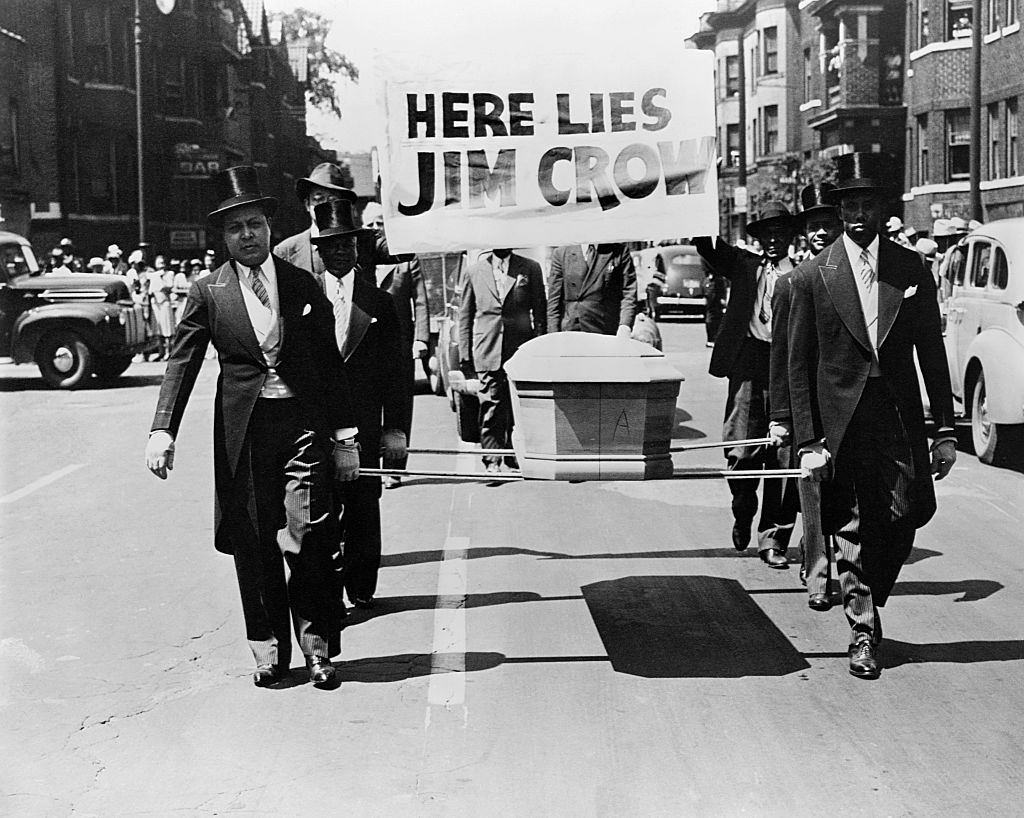 African-American men wearing tuxedos carry a coffin and a "Here Lies Jim Crow" sign down the middle of a street as a demonstration against "Jim Crow" segregation laws, 1944.