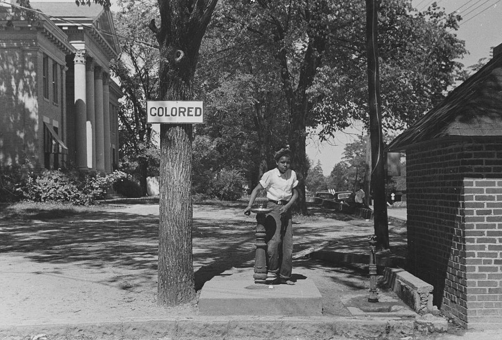 African American child using a water fountain, the tree next to the fountain has a sign attached that reads 'Colored' in order to enforce Jim Crow laws