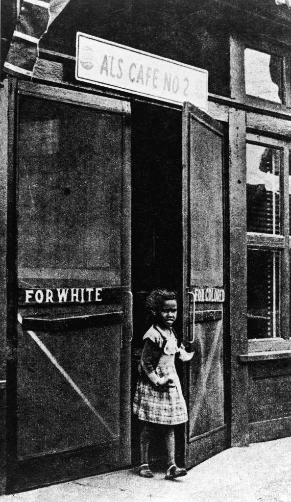 A black little girl leaves a cafe through a door marked 'For Colored,' circa 1950.