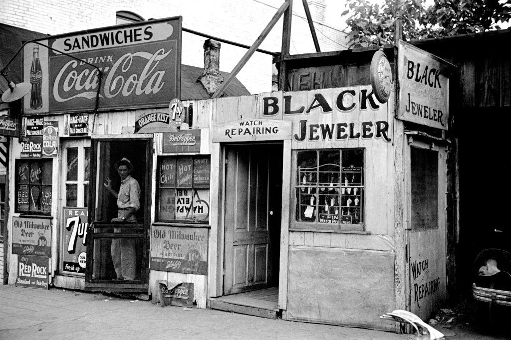 Shack-like Black Jeweler shop next to a small food store covered with beverage ads in a slum section of the city of Atlanta, 1938.