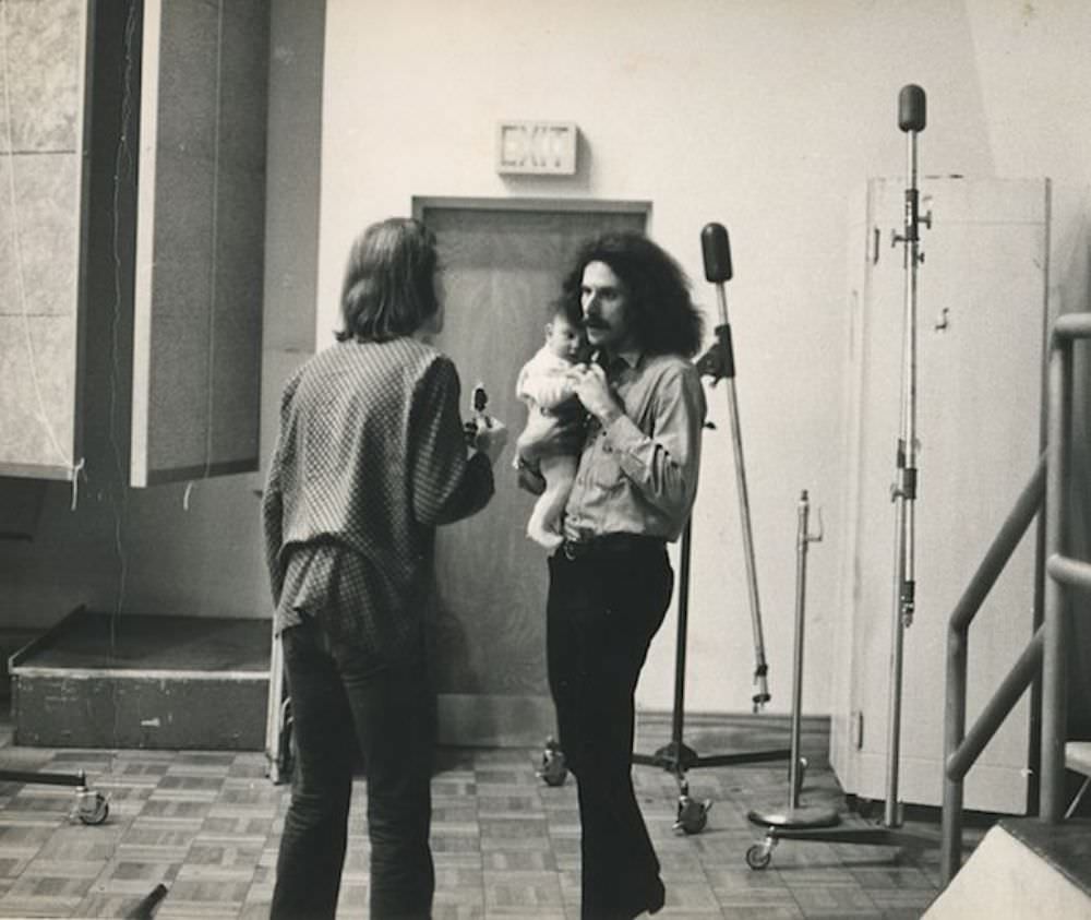 Photographer Bob Seidemann is seen holding a band member’s baby while talking with Big Brother’s road manager, David Richards..