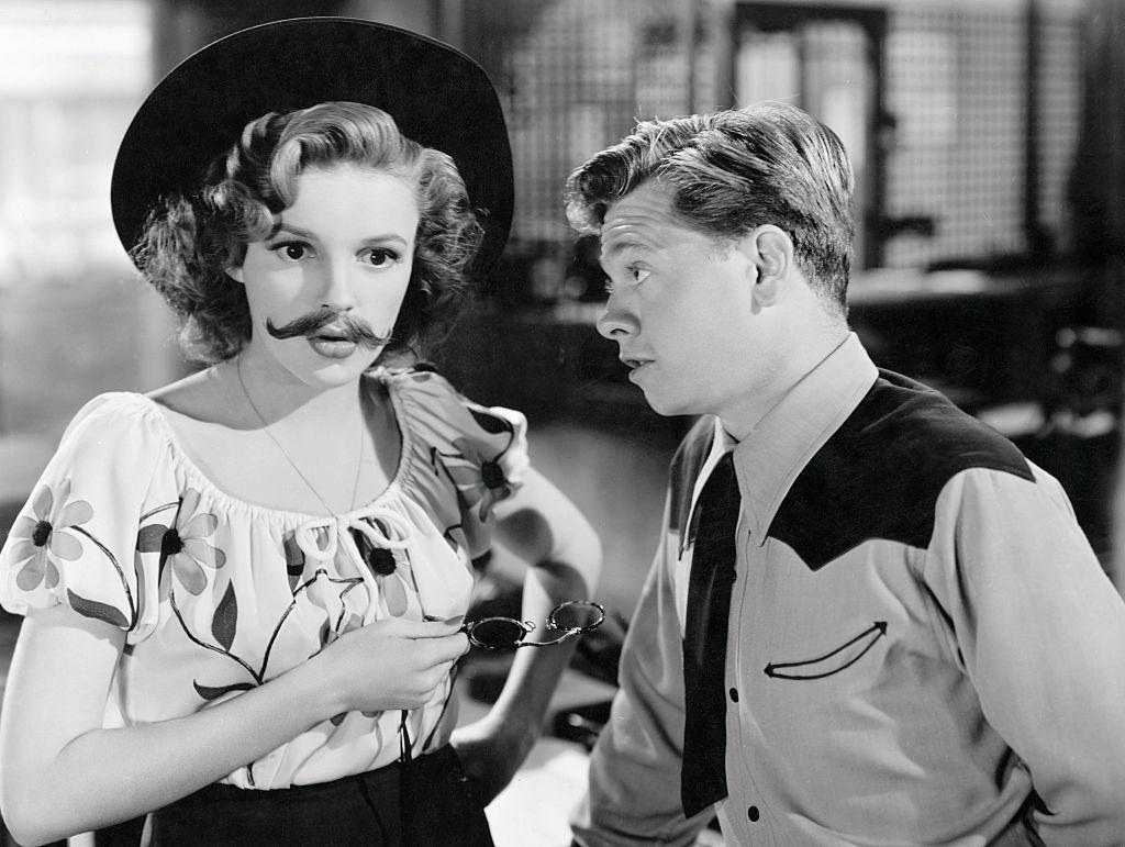 Mickey Rooney and mustachioed and be-hatted Judy Garland confer in the 1943 MGM musical comedy, Girl Crazy.