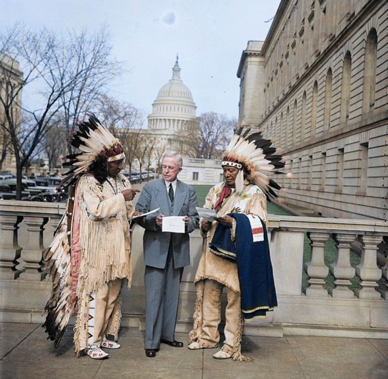 Two Native American elders, dressed in traditional garb discussing policy with a member of President Coolidge’s staff.