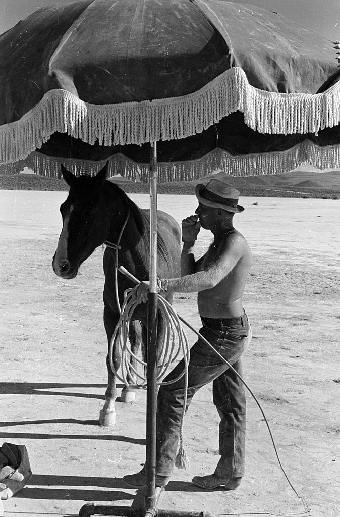 A trainer with a whip shares his shade with a horse on the set of John Huston's 'The Misfits'