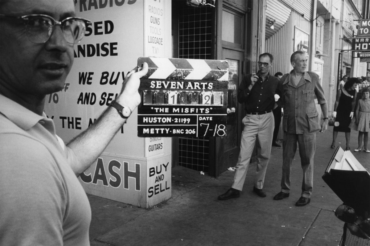 Clapperboard is marked scene 1 take 2 on the first day of shooting. Director John Huston and author Arthur Miller watching in the background.