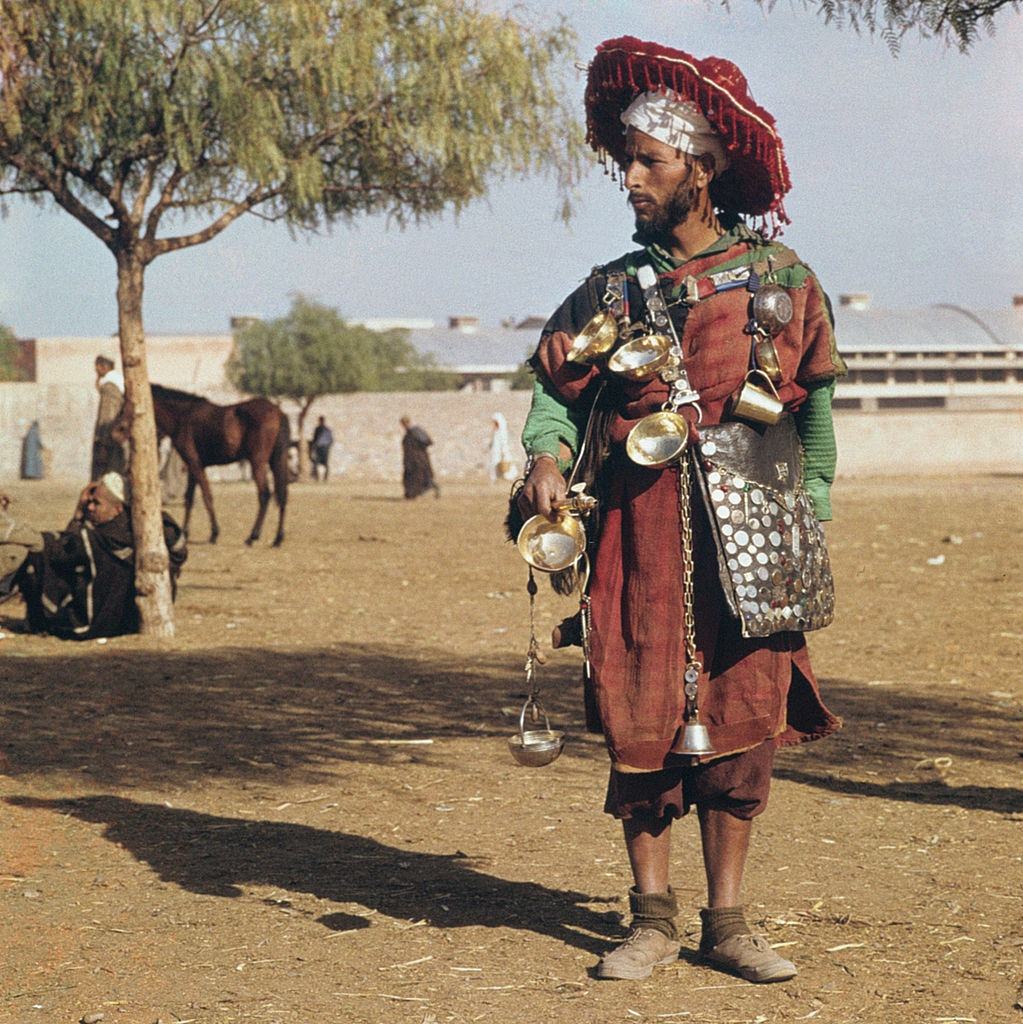 A peddler sells water in traditional clothes. Marrakech, 1960.