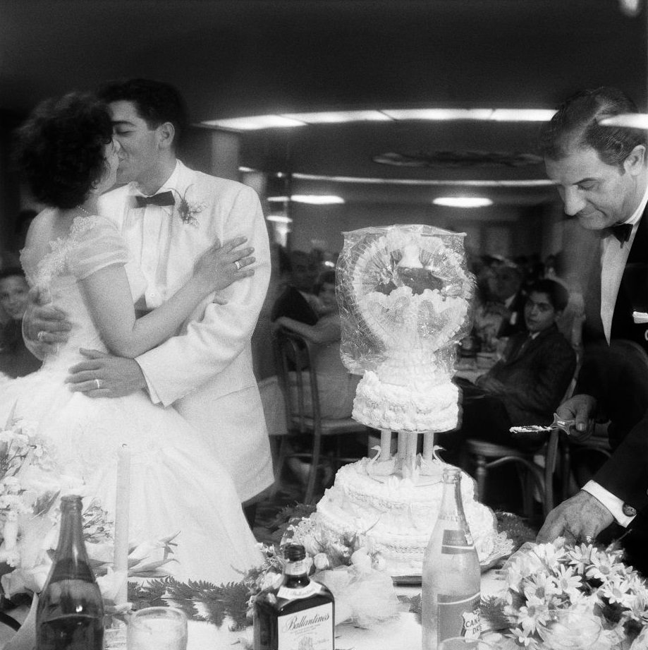 A newlywed couple kisses at their wedding reception, as their wedding cake is cut in Brooklyn Heights, March 1985.