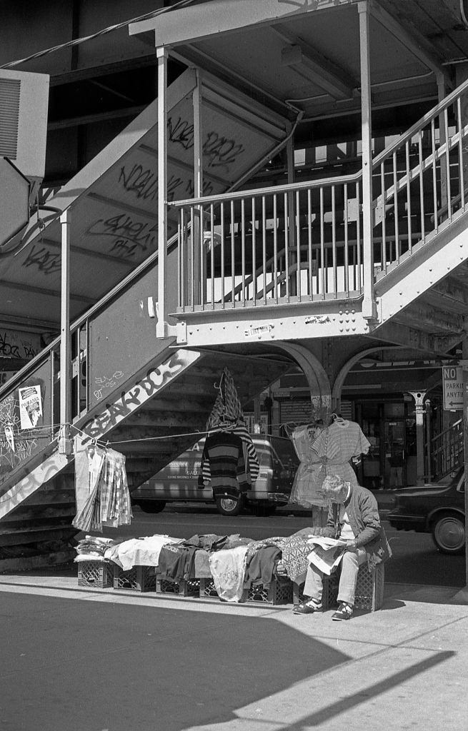 A vendor selling clothes next to the staircase leading to the 103rd Street elevated subway station in the Corona neighborhood. Queens, New York, 1990.