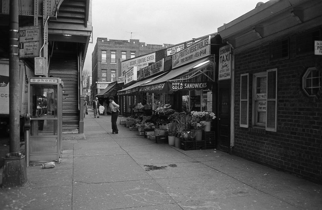 A lady exits a fruit and vegetable stand below the elevated subway line on Roosevelt Avenue in the heart of Corona, Queens. 5th November 1974