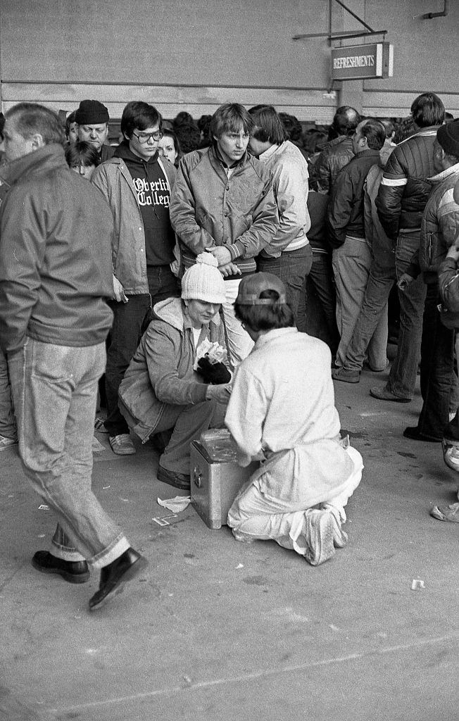 A woman crouches down to purchase a hot dog from a vendor during an opening day baseball game at Shea Stadium. Queens, New York, 1982