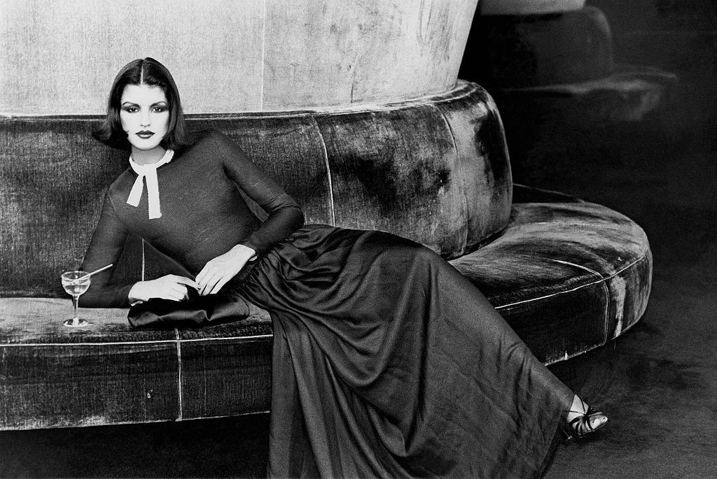 Janice Dickinson, seated on circular banquette with a cocktail drink. Vogue, 1975.