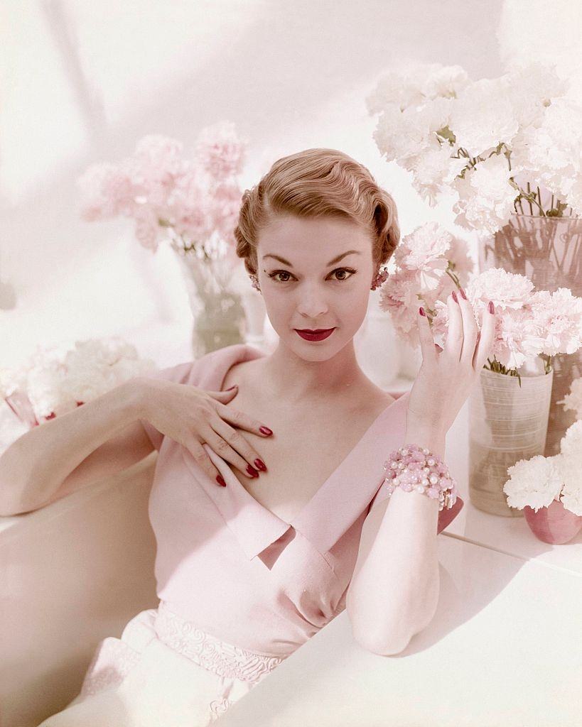 Jean Patchett seated among vases of pink and white carnations wearing pink open neck linen dress with wide lapel collar and matching white with pink brocade skirt. Vogue 1950