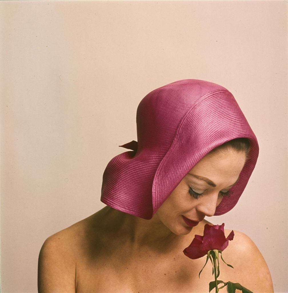 Jean Patchett in a pink hat, looks down at a pink rose, 1961.