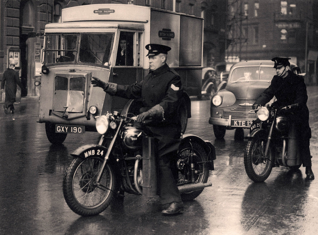 Two Manchester City Police officers pull their BSA motorcycles out into traffic on a wet day in Manchester city centre, 1956