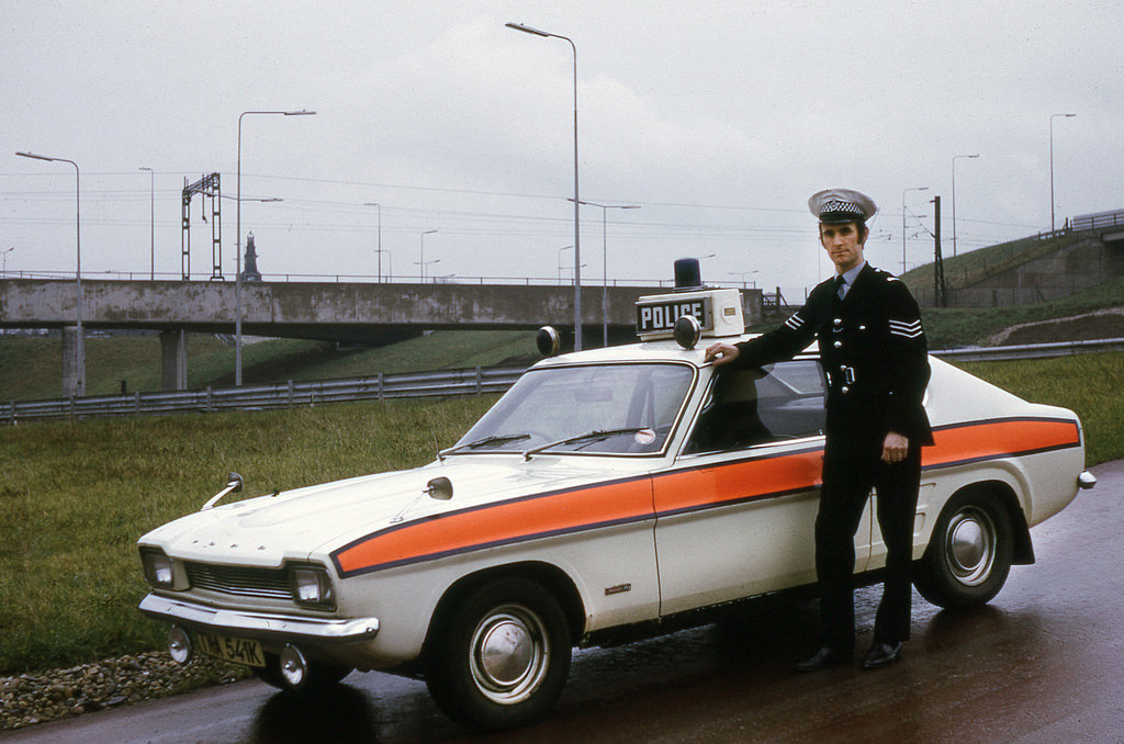A police sergeant stands beside a classic police vehicle of the era in February 1975
