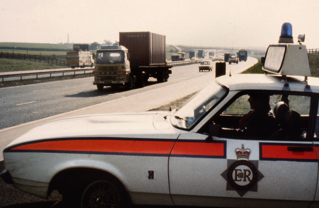 A Ford Capri sits beside a motorway in the region in the 1980s