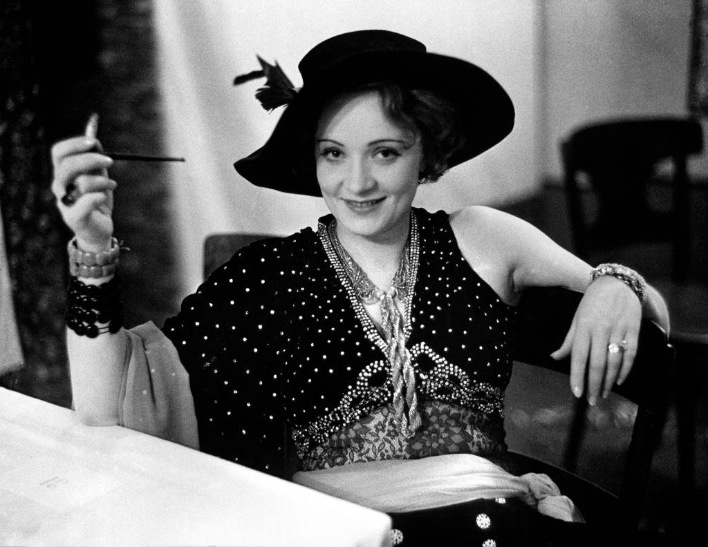 Marlene Dietrich in evening dress and hat, smiling while sitting at table alone during Pierre Ball.