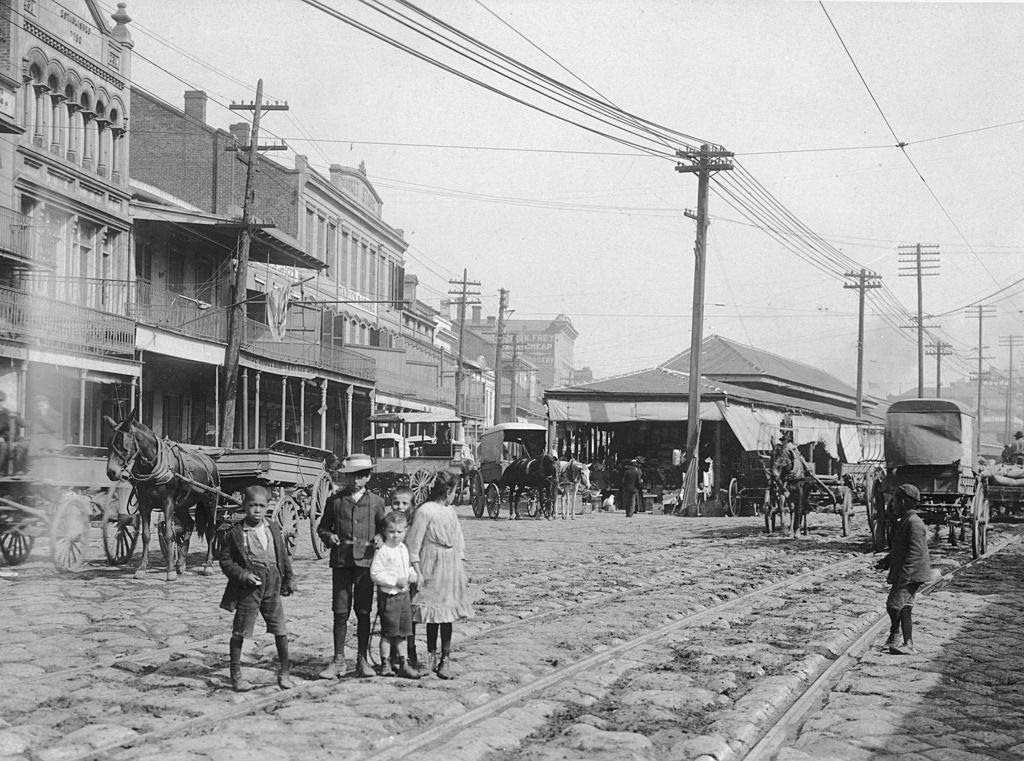 A group of children stand along a carriage track in a cobblestone street in front of French Market. New Orleans, 1895.