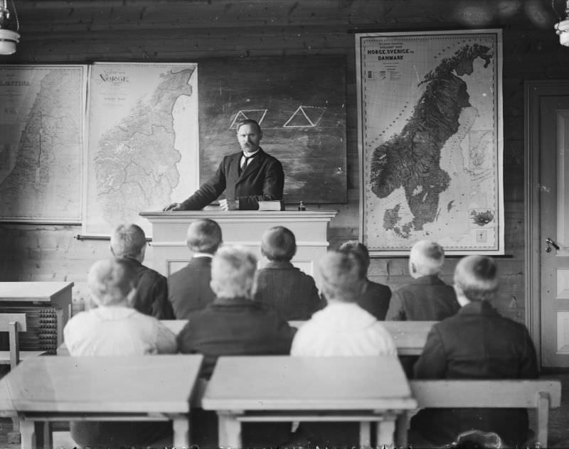 The school class seems to boys only. Maps of Norway and Scandinavia are hanging on the wall behind the teacher