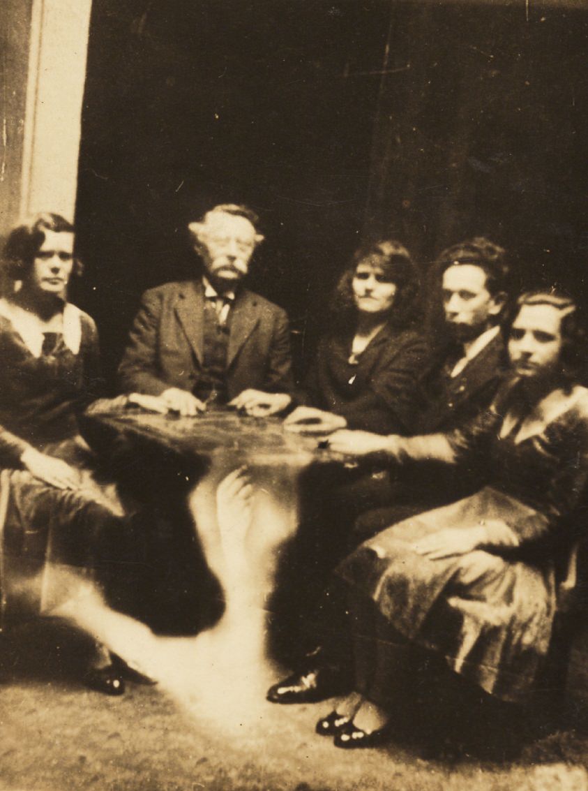 A seance with a ghostly arm superimposed under the table.