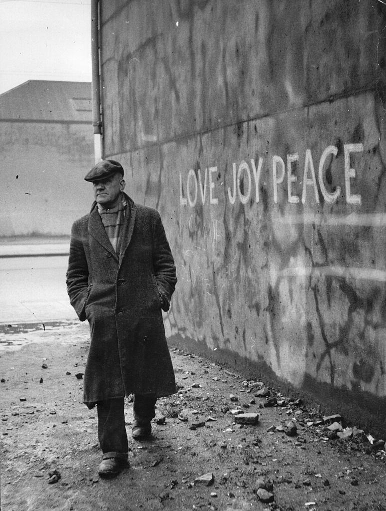 Retired lion-tamer Alec Robinson walks past a Belfast wall scrawled with the slogan 'Love - Joy - Peace', 1955.