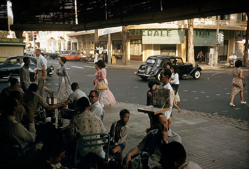 Male patrons of a sidewalk cafe check out women pedestrians in Saigon, 1961