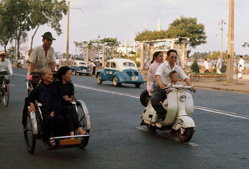 A man drives his wife and son on a scooter past passengers on a cyclo in Saigon, 1961