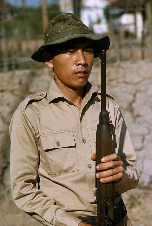 A stern-faced Popular Forces soldier stands at attention with his gun in Saigon, 1965