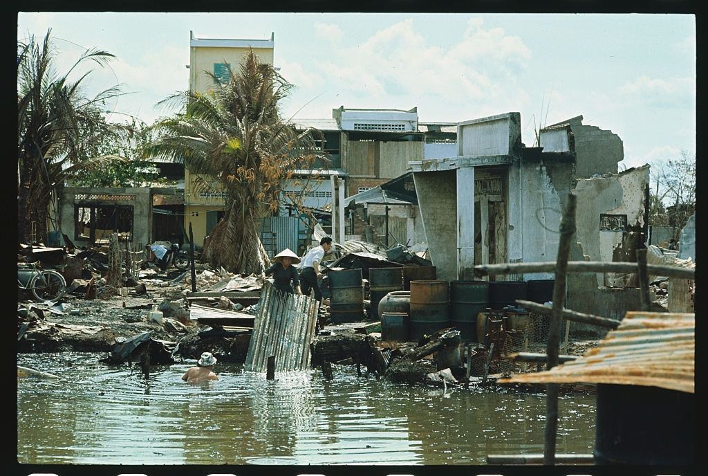 Vietnamese woman takes a short rest as her husband working in the water looks around for salvaged tins and other material to be used in rebuilding their home, 1960.