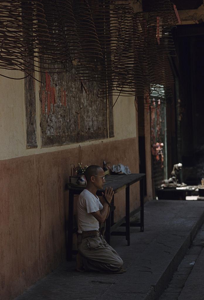 A man kneeling on a sidewalk, prays with folded hands, holding a Buddhist Mâlâ, 1961.