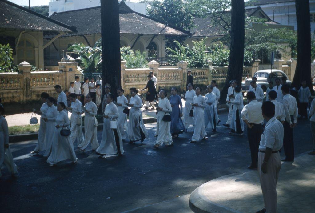 Female mourners, wearing white ao dai, walking on a tree-lined street during Colonel Hoang Thuy Nam's funeral procession, Saigon, 1961.