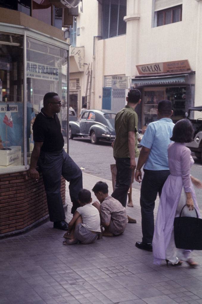 In a street in Saigon, two Vietnamese children shine the shoes of a black man, in front of the window of an Air France agency.