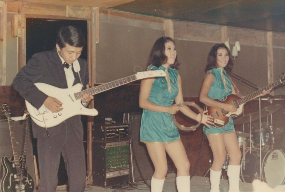 A man playing a white electric guitar and two young women, wearing turquoise minidresses, playing the tambourine and another guitar, performing on a stage, Vietnam, 1965.