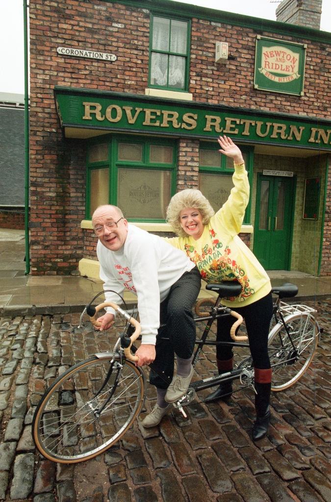 Julie Goodyear and Roy Barraclough pictured on a tandem bike on the set of Coronation Street, 13th February 1990.