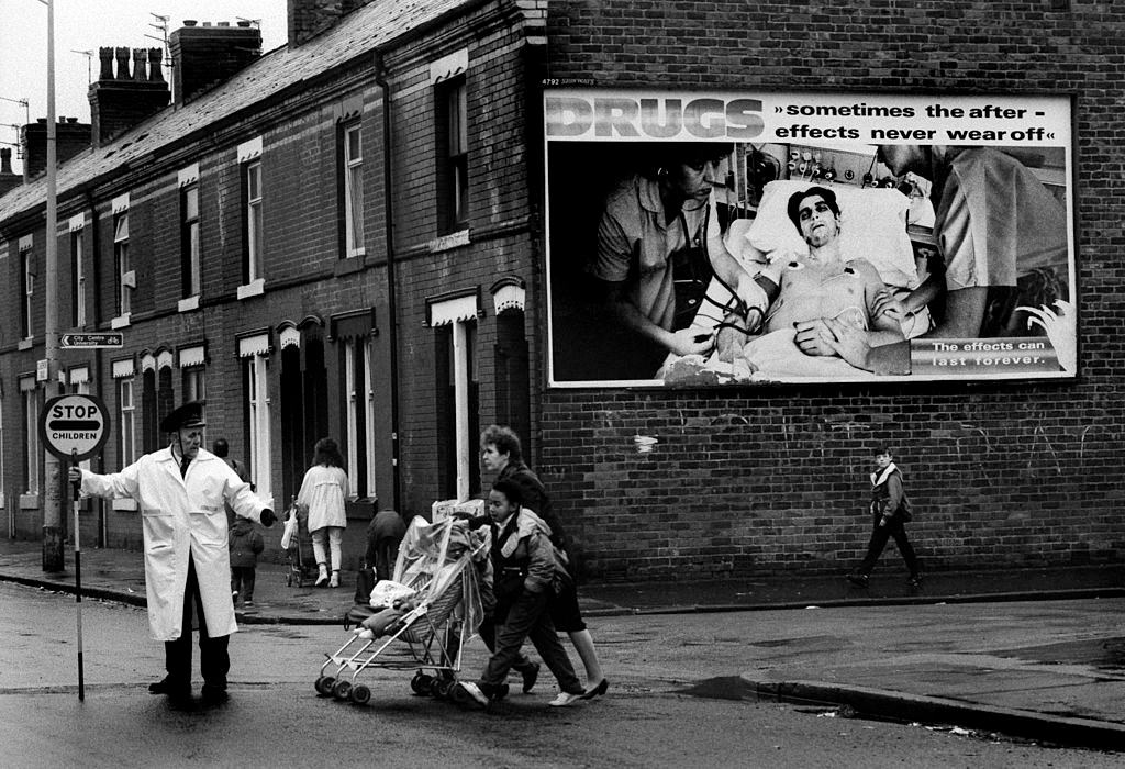 A lollipop man guides children to school across a road in Moss Side, an inner city district of Manchester, 1990.