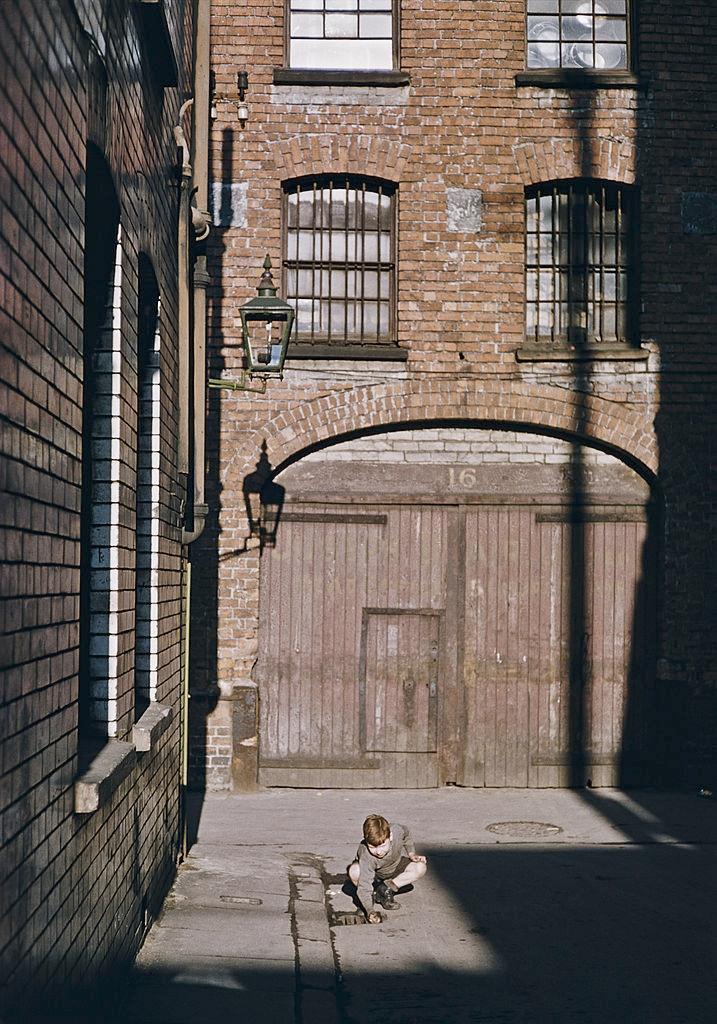 A boy crouching by a drain in a courtyard, Belfast, 1955.