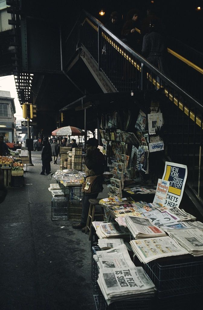In Brooklyn, a black newspaper seller near his stall under a subway staircase, 1973.