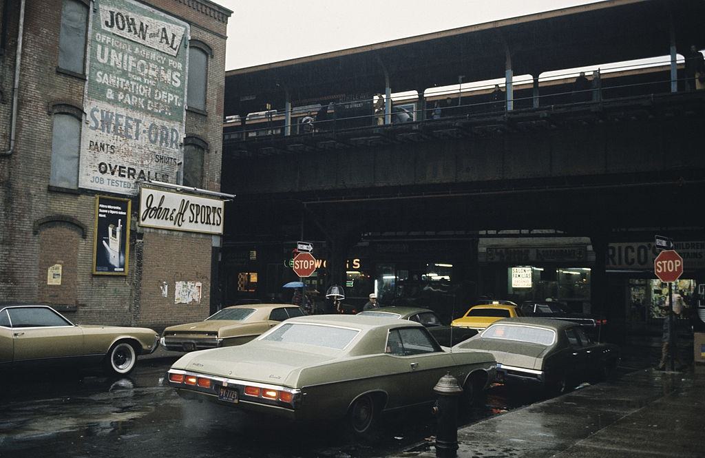 A street with parked cars, passing under a subway traffic bridge, Brooklyn, 1973.
