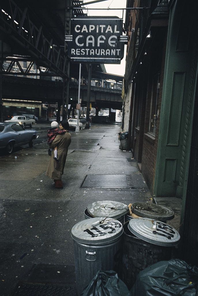 A street overhung by bridges, on a sidewalk, near the front of a cafe, Brooklyn, 1973.